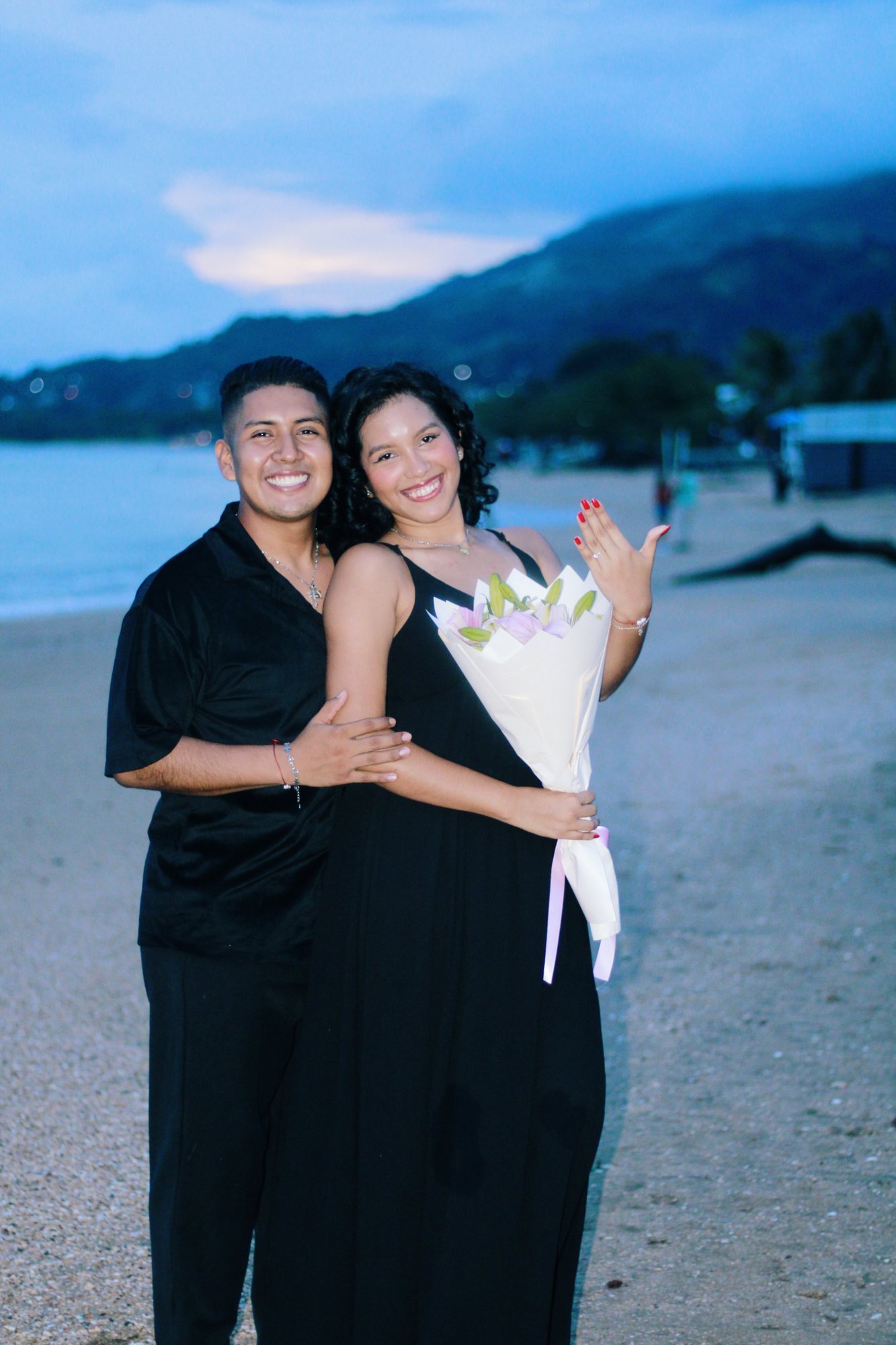 Pareja sonriente mostrando el anillo de compromiso y un ramo de flores